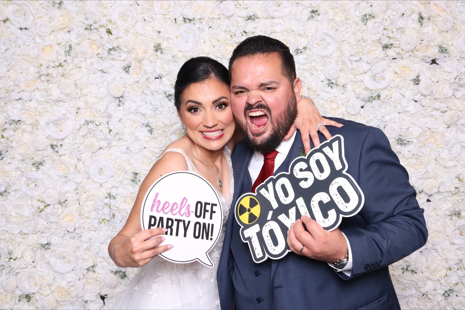 A bride and groom laughing with props in front of a white flower wall at their wedding reception