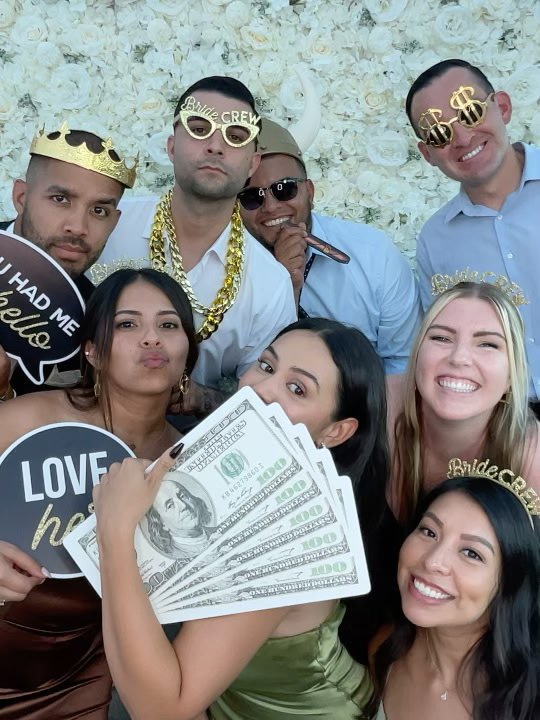 A group of guests posing together with crowns, sunglasses and prop signs in front of a white flower wall